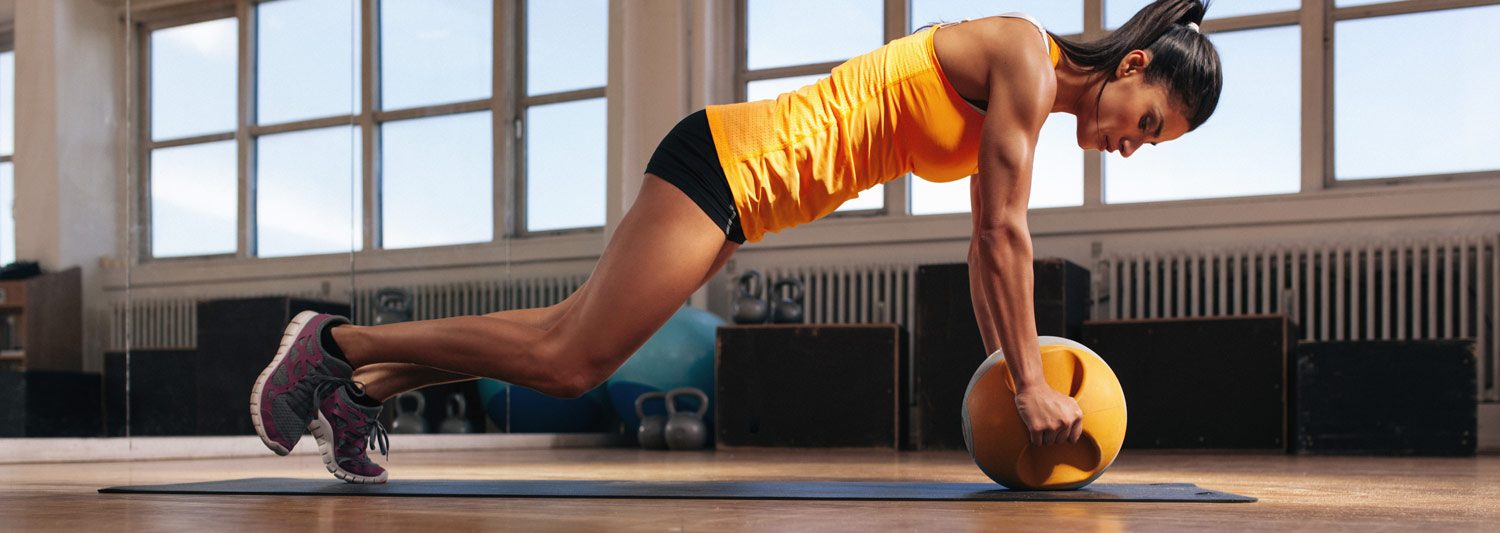 Woman in a yellow shirt and black shorts doing a gentle pushup while resting hands on a kettleball.